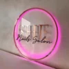 Elegant circular acrylic neon sign with a soft pink glow, featuring the "SHE Nails Salon" branding in a stylish and modern design.