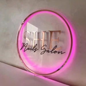 Elegant circular acrylic neon sign with a soft pink glow, featuring the "SHE Nails Salon" branding in a stylish and modern design.