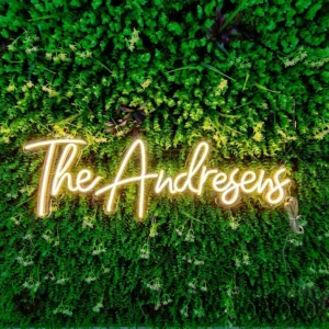 The Family Name Neon Sign from the Custom Wedding Stylish Personalized Décor collection glows warmly with The Andresens against lush green foliage, capturing the charm of a personalized family name sign.