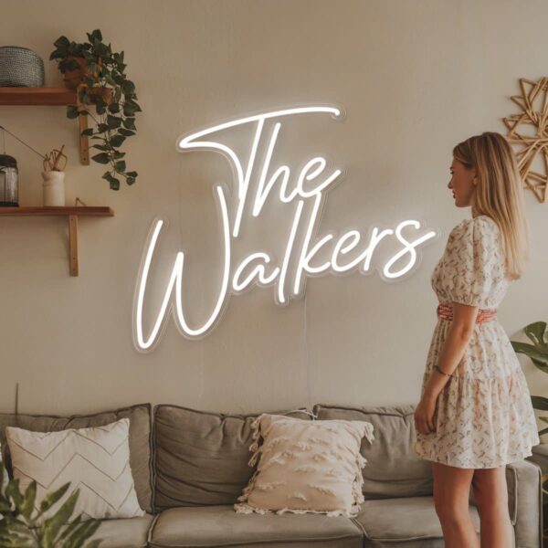 A woman admires The Walkers on a Family Name Neon Sign - Custom Wedding Stylish Personalized Décor in the living room, wearing a floral dress. Decorative pillows line the sofa under it, while lush plants and stylish wall decor enhance the cozy atmosphere.