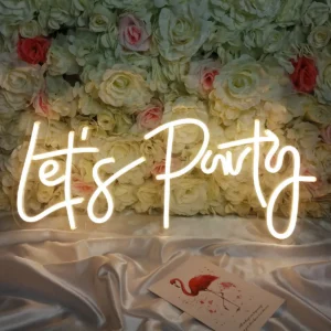 A Let’s Party Neon Sign - Fun & Vibrant Event decor illuminates a backdrop of white and pink roses with a warm glow, while a flamingo design card rests elegantly on silky, light-colored fabric.