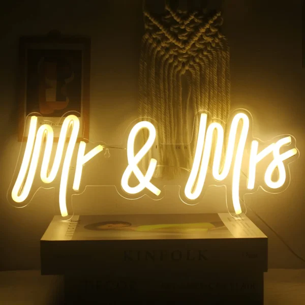 The CustomElegant Mr. & Mrs. Neon Sign casts a warm yellow glow in cursive, illuminating a wall with macramé decor and books below.