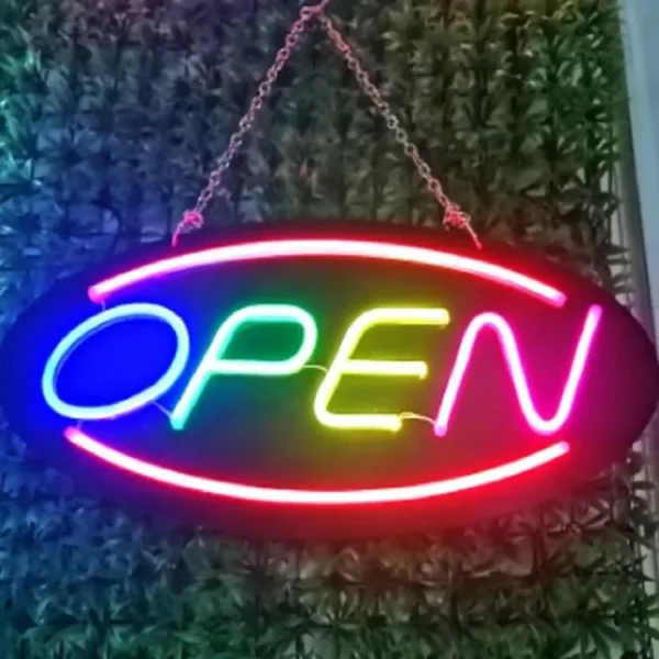 The Neon Open Sign, an energy-efficient LED display for businesses, shines in vivid red, blue, yellow, and green. Oval-shaped and chain-suspended, it adds a vibrant touch against a leafy green backdrop.