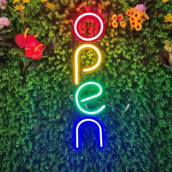 The vibrant Neon Open Sign – Eye-Catching, Custom Energy-Efficient LED Design for Businesses is mounted vertically on a lush green wall with colorful flowers.