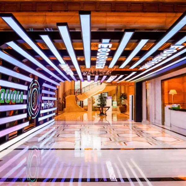 A modern interior with horizontal light panels on the ceiling features a staircase in the background and a glossy geometric floor. A central plant enhances the space, while a custom Full Color LED Digital Display Sign adds vibrant ambiance with WiFi & USB programmable features for dynamic content display.