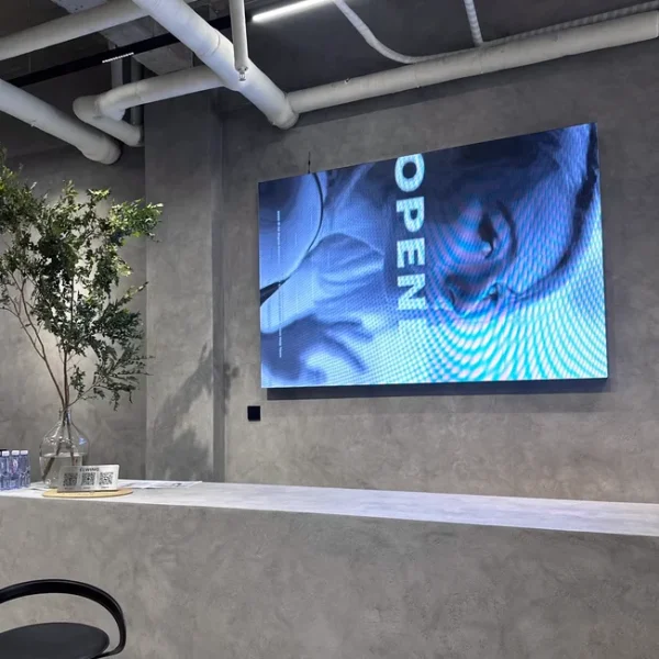 A modern interior features a minimalist counter and tall potted plant. A Full Color LED Digital Display Sign, mounted on the textured grey wall, shows a blue OPEN image.