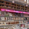 A store shelf with model kits and puzzles sits under a Full Color LED Digital Display Sign. A banner saying Merry Christmas in red, with festive graphics, hangs above. Potted plants are placed atop the shelves.