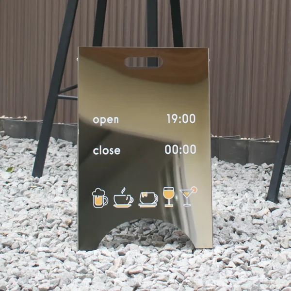 A Custom Sandwich Board Metal A-Frame Sign on a gravel path features drink icons, indicating opening at 19:00 and closing at 00:00.