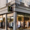 A corner view of a clothing store showcases large glass windows with neatly arranged garments on racks. Above the entrance, an energy-efficient solar sign reads TribalFit, using Solar Signs Light Boxes with double-sided illumination & weatherproof outdoor signage