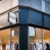 A clothing store with a big window display showcases garments on hangers. The GLOW ESSENCE name shines brightly in Solar Signs Light Boxes above the entrance, surrounded by modern black framing and lush greenery at this chic boutique.