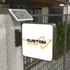 A Solar Signs Light Box, ingeniously designed as a modern solar-powered square outdoor sign, reads CUSTOM YOU SIGN with an orange and black curved arrow graphic encircling the text, set against a stone wall and lush greenery while attached to a metal fence.