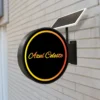 A round, black solar sign in cursive is mounted on a brick wall, featuring vibrant yellow and orange accent rings. The Solar Signs Light Box offers energy-efficient double-sided illumination and is weatherproof for outdoor use, powered by the solar panel on top.