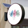 Round outdoor solar sign. It has a small solar panel on its bracket attached to a wooden wall. Product: Solar Signs Light Boxes - Energy-Efficient Double-Sided Illumination & Weatherproof Outdoor Signage.
